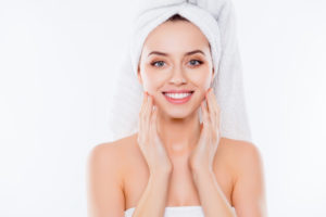 Portrait of stunning woman in towel and turban on head enjoying touching her perfect face skin with fingers isolated on white background